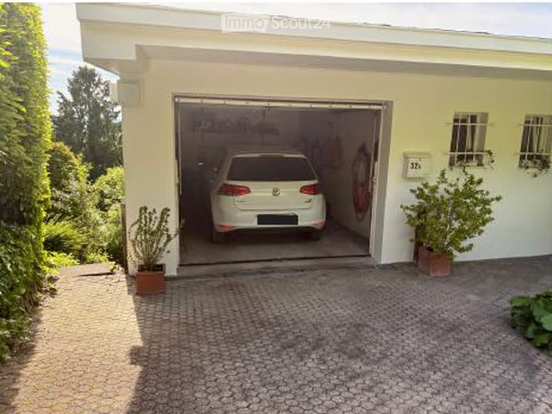 A modern single-story house with a large garage door open, revealing a parked car inside. The driveway is paved with cobblestones, and there are potted plants on either side of the entrance.