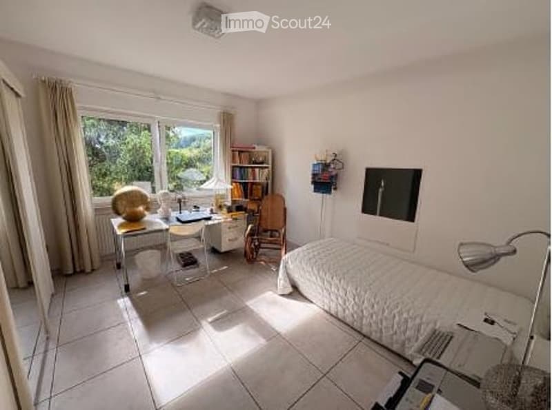 Bright bedroom with large window offering a scenic view, featuring a single bed, desk with chair, bookshelf, and a lamp.