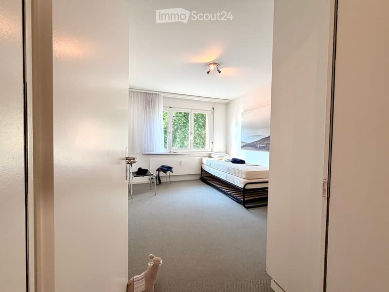 A small, well-lit bedroom with a single bed, two stools, and a picture frame on the wall. The room has large windows with white curtains and a carpeted floor.
