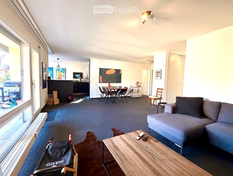 Open-plan living and dining area with a grey sofa, wooden coffee table, dining table with chairs, and a bar counter with a fridge. Large windows provide natural light.