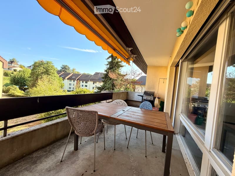 Balcony with a wooden table, four chairs, and a grill, featuring an orange awning and a view of the neighborhood.