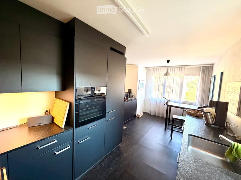 Modern kitchen with dark grey cabinets, stainless steel appliances, and a central island with a sink and coffee maker. The space includes a small dining area with a table and stool, and a window with curtains letting in natural light.