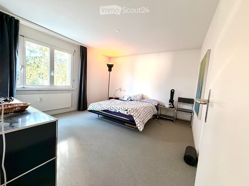 Modern bedroom with a large bed, nightstand, chair, floor lamp, and a painting on the wall. The room features large windows with dark curtains and a carpeted floor.