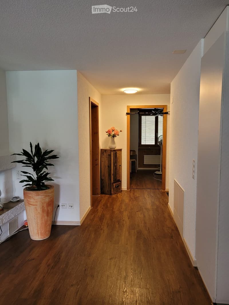 A well-lit hallway with wooden flooring, white walls, and a large potted plant on the left side. A small wooden cabinet with a vase of flowers is visible further down the hallway.