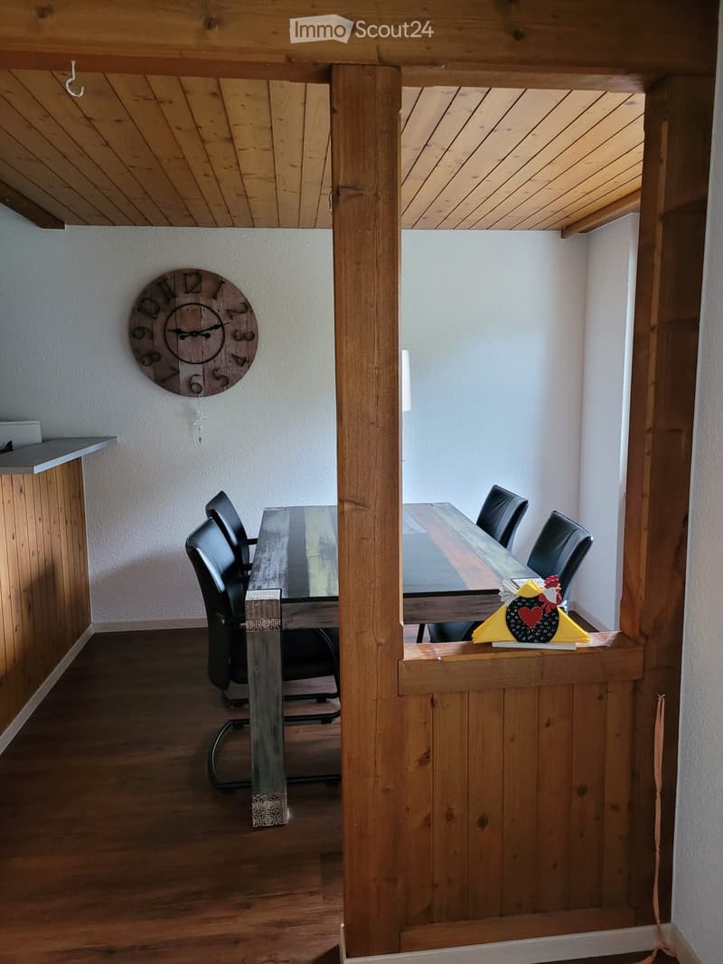 Open-plan dining and lounge area with wooden beams, a round wooden clock, a glass dining table, and black chairs.