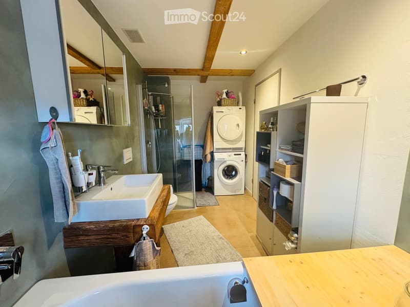 Modern bathroom with a sink, bathtub, shower, washing machine, and wooden cabinets. Features exposed wooden beams and a light-colored wooden floor.