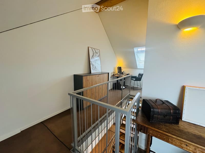 A modern, well-lit room with wooden flooring, a staircase with a metal railing, a desk with a computer, a chair, a cabinet, and a small table with a suitcase and picture frame.