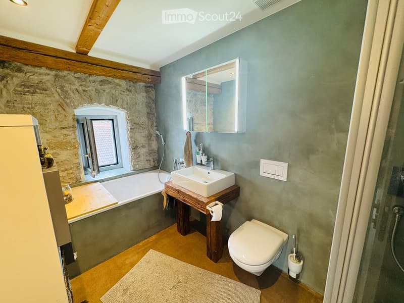 Bathroom with a bathtub, sink, toilet, mirror, and a window. The walls are painted in a muted green color, and the floor is wooden.