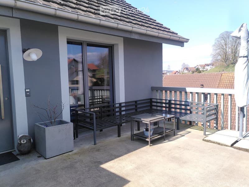 Balcony with metal furniture including a table, chairs, and a bench, overlooking a residential area with tiled roofs.