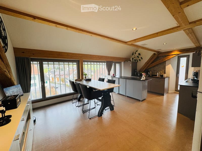 Bright, open-plan living and dining area with exposed wooden beams, large windows, modern kitchen, dining table with chairs, and decorative items.