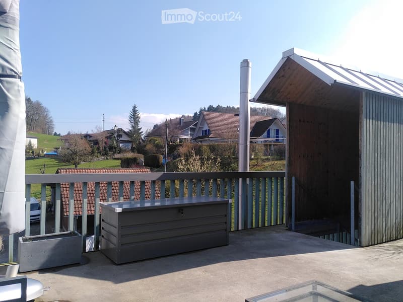 Balcony with metal railing, storage box, planter, and a view of a residential area with houses and greenery.