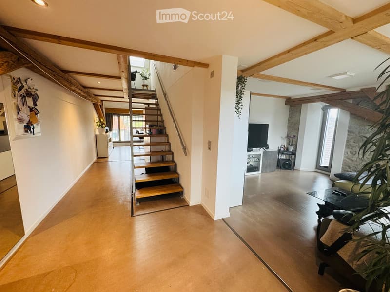 Spacious hallway with wooden staircase leading to upper level, exposed wooden beams, modern lighting, and open-plan living area visible at the end.