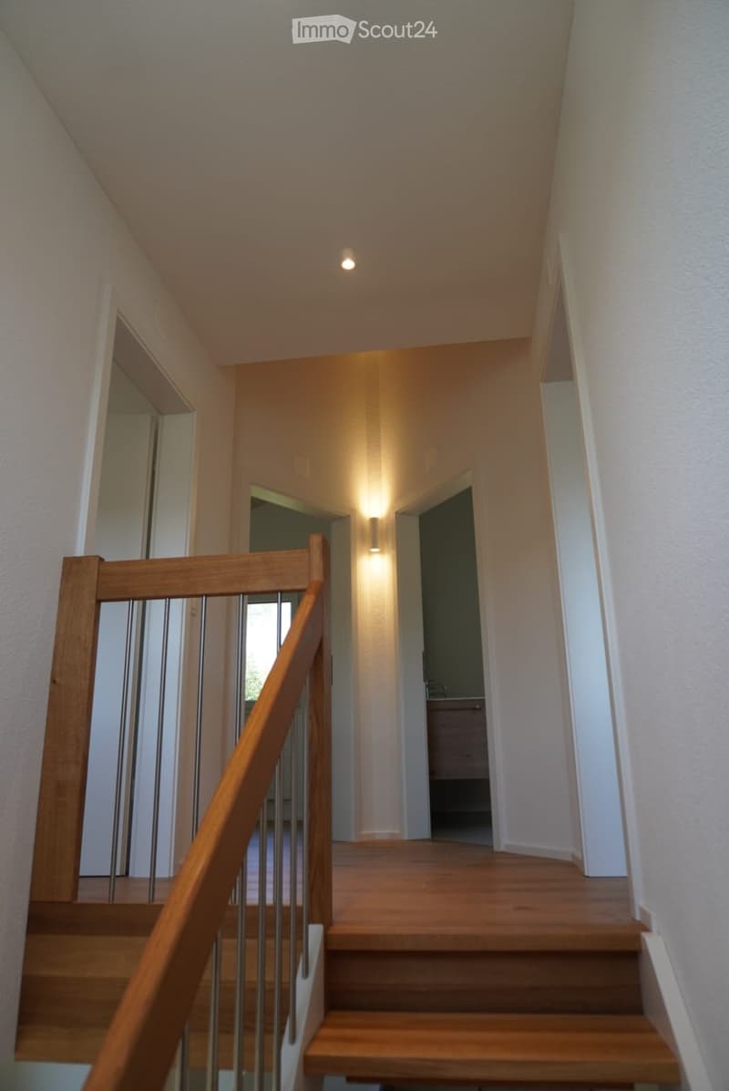 Wooden staircase with metal railing, beige walls, and recessed lighting. Open doorways lead to adjacent rooms.
