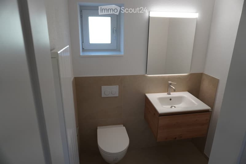 Modern bathroom with a toilet, sink, and mirror. The sink has a sleek wooden cabinet and a modern faucet. The walls are painted in neutral tones with a beige lower section.