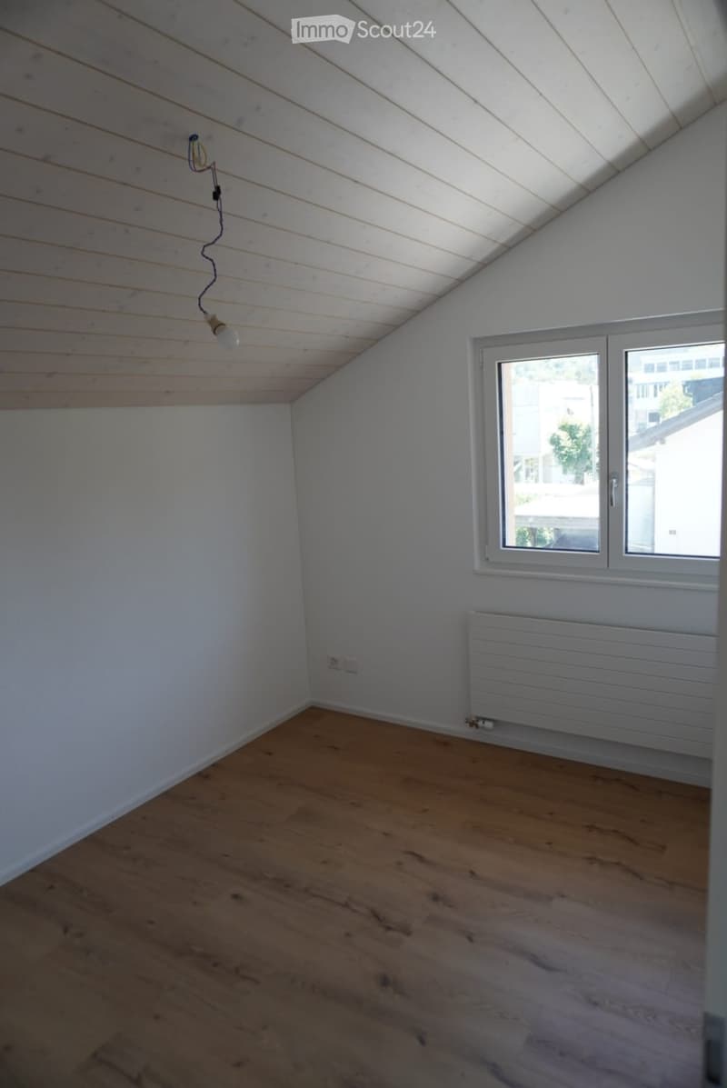 Empty attic room with wooden flooring, white walls, and a slanted wooden ceiling. Features a single window with white frame and a radiator below it.
