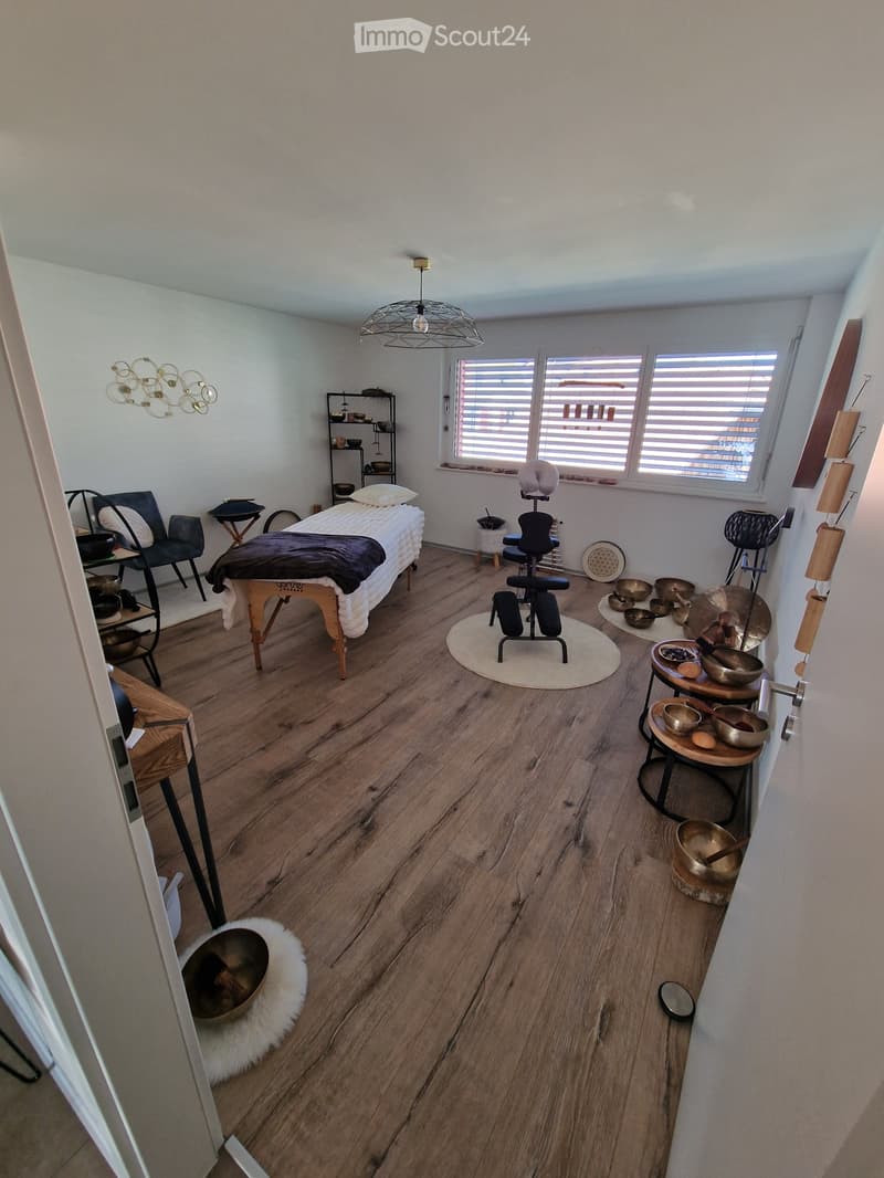 A well-lit room with wooden flooring, a massage table, chairs, a small table with bowls, and a shelf with decorative items.