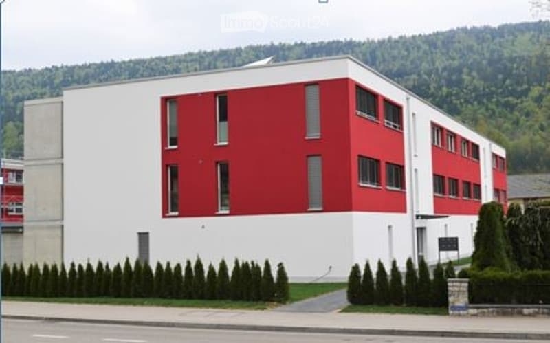 Modern two-story building with white and red facade, multiple windows, and neatly trimmed hedges in front.