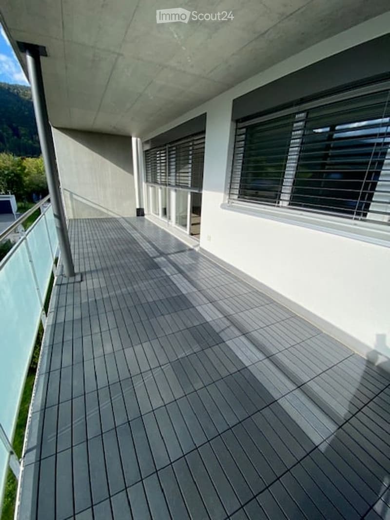 Modern balcony with grey tiled flooring, white walls, large windows with blinds, and a view of the surrounding landscape.