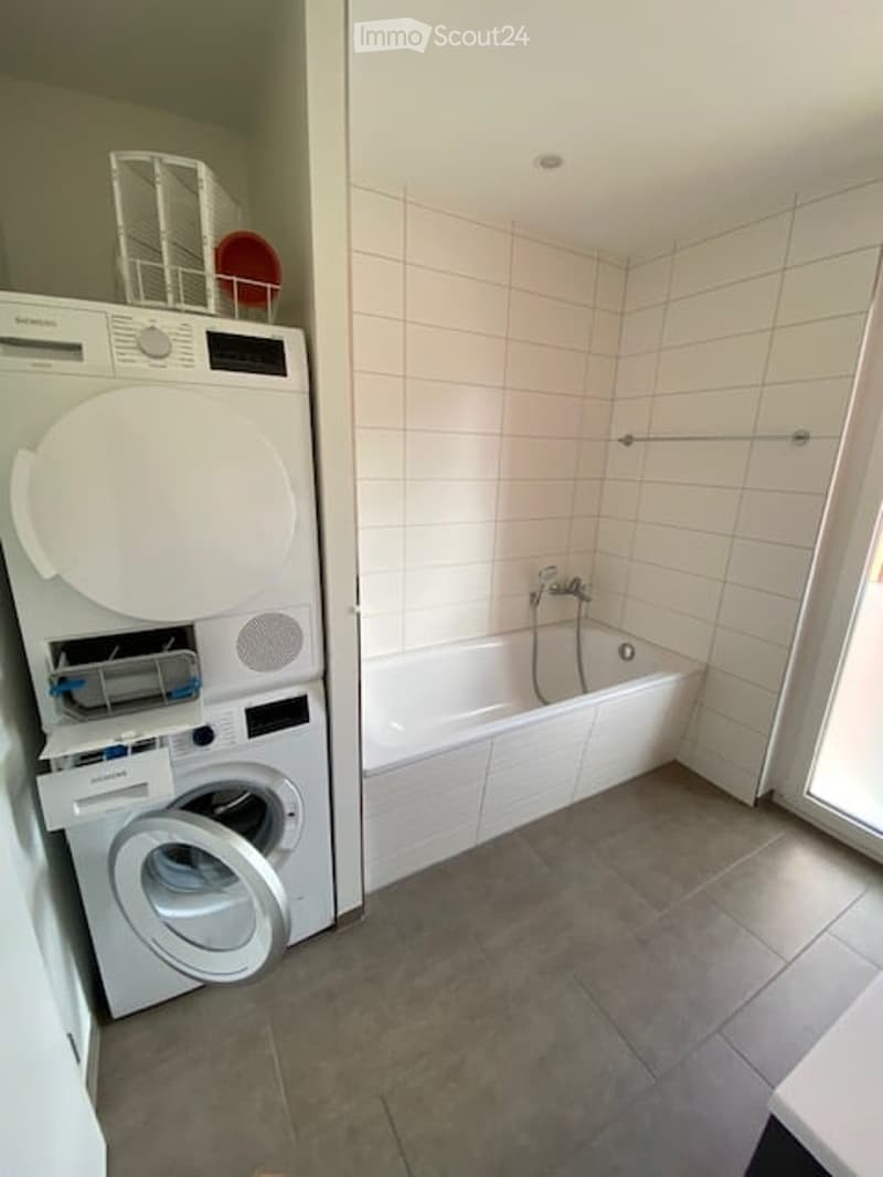 Modern bathroom with a white tiled shower area, a bathtub, and a stacked washing machine/dryer unit. The floor is tiled, and there is a towel rack visible.