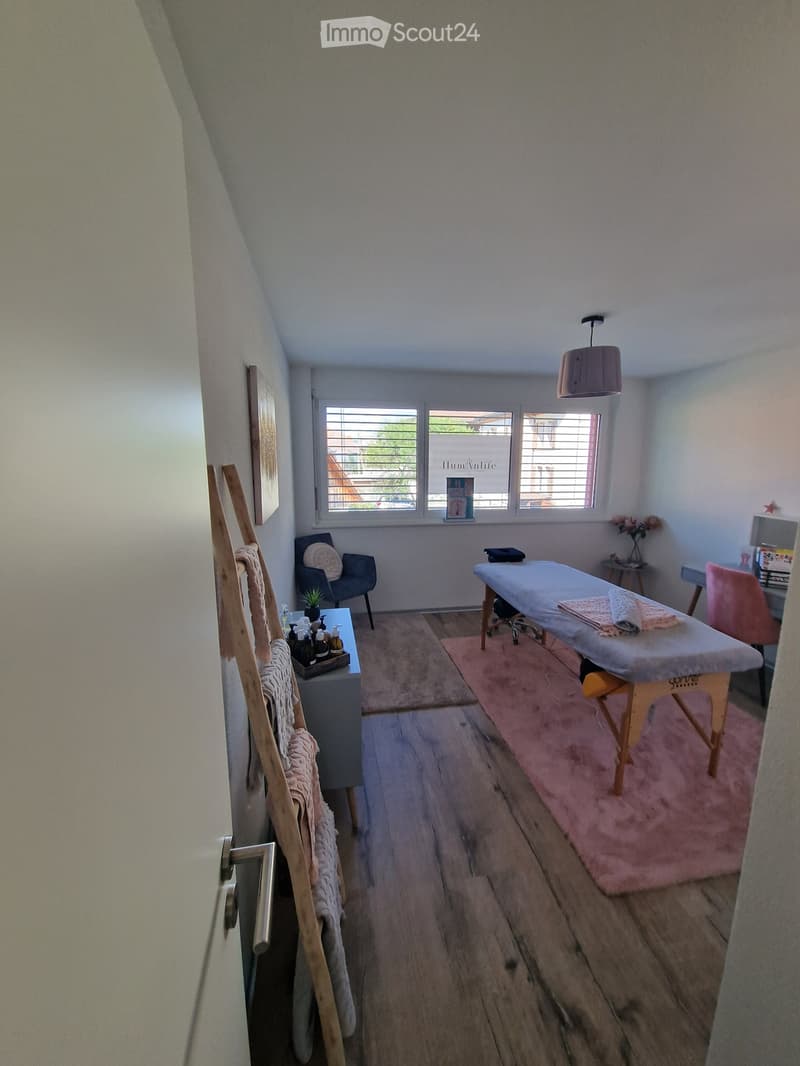 A well-lit room with wooden flooring, a massage table, a small wooden shelf with bottles, a chair, and a lamp.