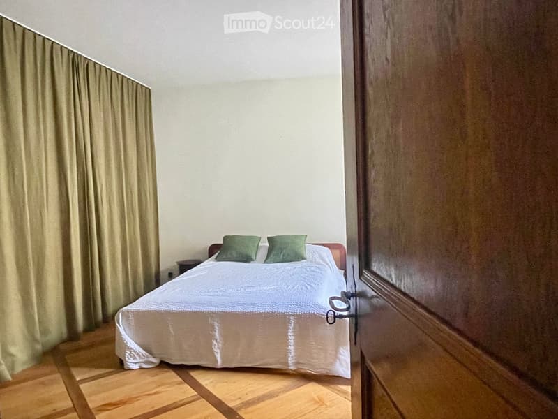 A small bedroom with a neatly made bed, two green pillows, and a nightstand. The room features wooden flooring and light-colored walls.