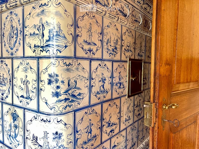 A narrow hallway with intricately designed blue and white tile panels featuring historical and scenic artwork, leading to a wooden door with a brass handle.
