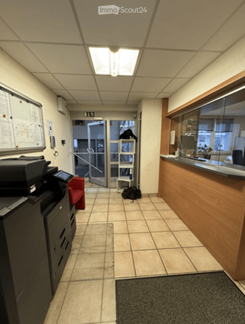 Empty hallway with tiled floor, white walls, and a reception area visible at the end. There is a printer, a chair, and a small table with items on it.