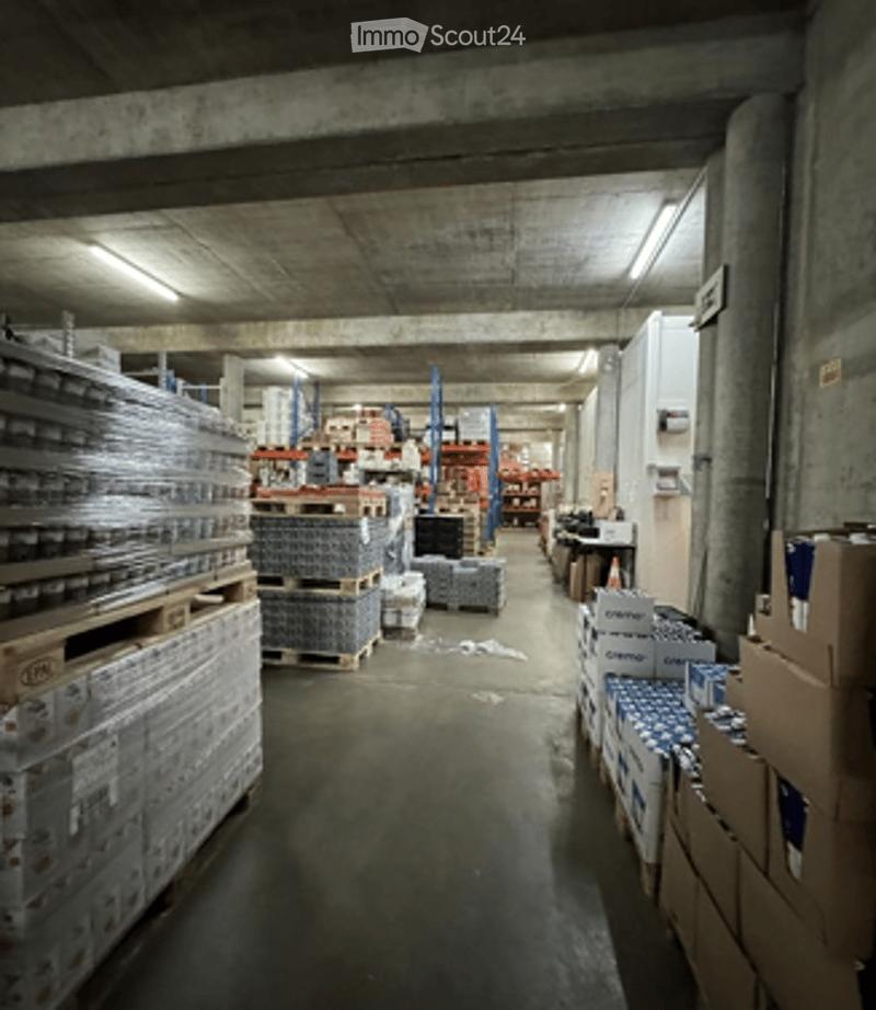 Warehouse space with concrete floors and ceilings, filled with pallets of boxes and shelves stocked with various items. Worktable and storage units visible along the sides.