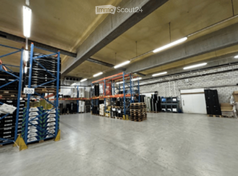 Large industrial-style warehouse with high ceilings, exposed beams, and bright lighting. Shelves, pallets, and storage units are visible, along with a computer setup in the background.