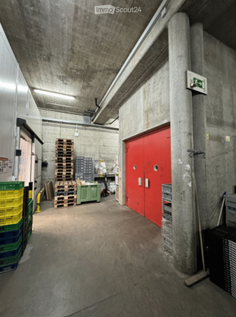 Empty warehouse or storage area with concrete walls and floor, large red garage door, stacked pallets, and storage crates.