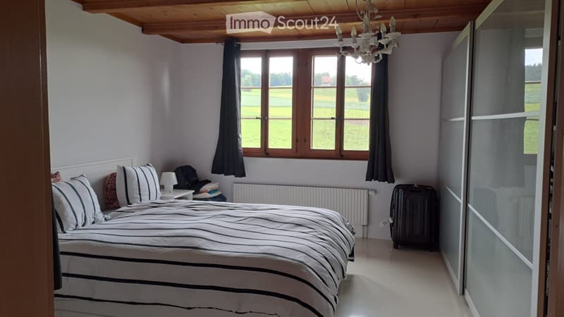 Cozy bedroom with a wooden ceiling, large window with black curtains, bed with striped bedding, nightstand with lamp, and a wardrobe with mirrored doors.