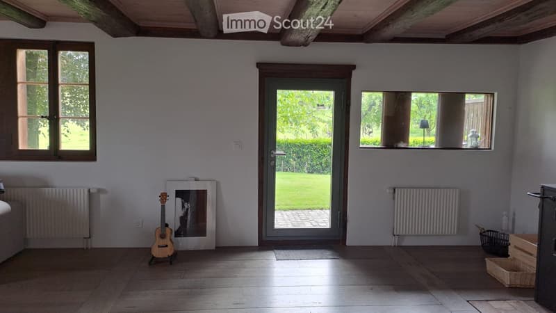 Bright, unfurnished room with wooden flooring, large windows, and a door leading to a garden. A guitar and a picture frame are placed against the wall.