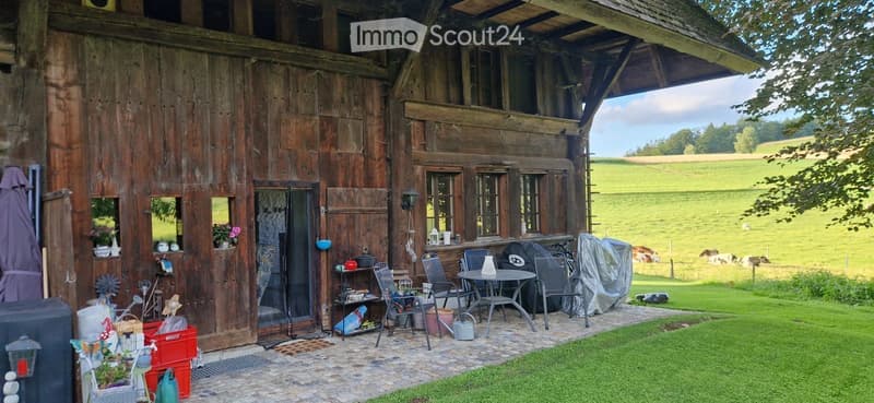 A rustic wooden barn with a covered outdoor seating area, featuring a table and chairs, surrounded by a green lawn and a scenic countryside view.