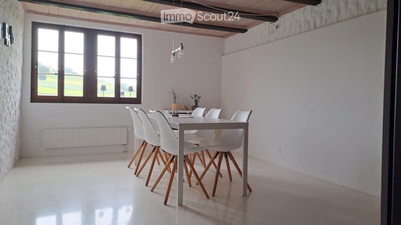 Bright dining room with white walls, large window, modern dining table and chairs, and a small flower vase on the table.