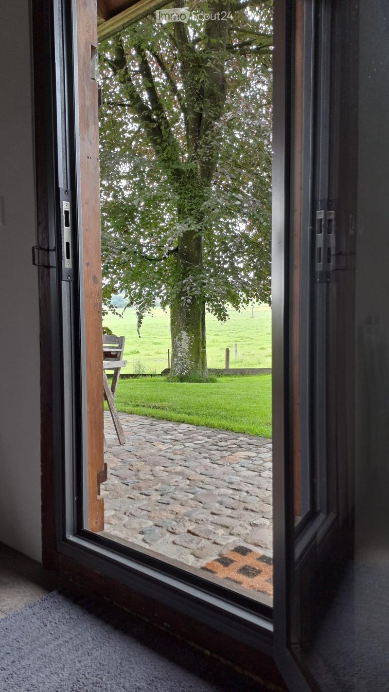 Open wooden-framed door leading to a stone-paved patio with a view of a large tree and green field.
