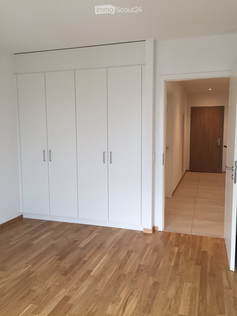 Empty hallway with light wooden flooring and white built-in wardrobes on one side, leading to another room with a wooden door.