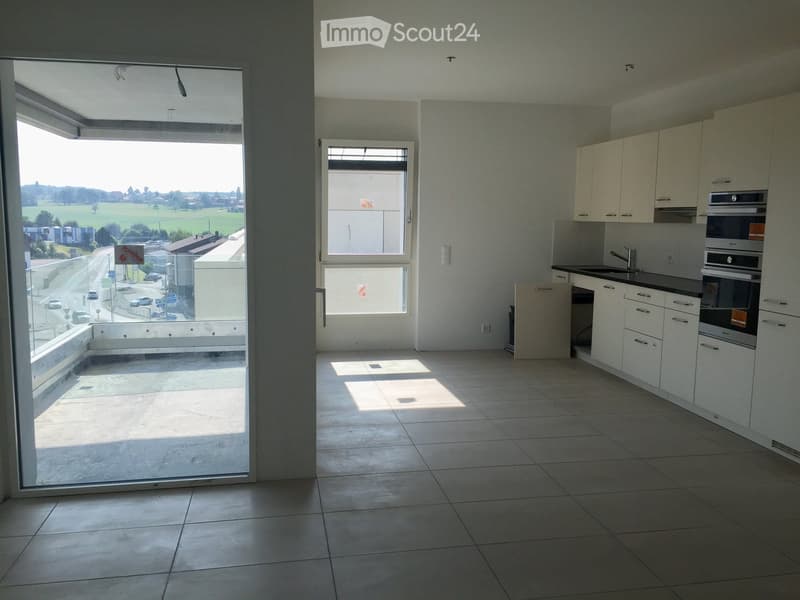 Empty kitchen with white cabinets, tiled floor, and large windows, connected to a balcony overlooking a road and landscape.