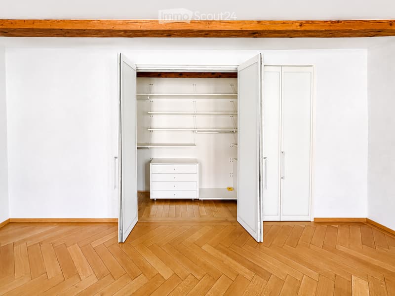 Empty room with light wooden herringbone flooring, white walls, and an open closet with white shelves and a small white dresser inside.