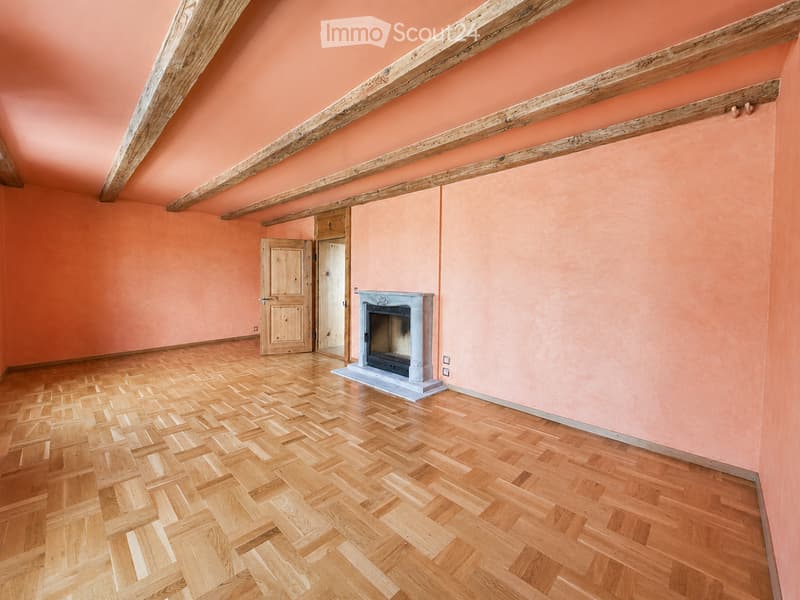Empty room with peach-colored walls, wooden herringbone flooring, exposed wooden beams on the ceiling, and a fireplace with a stone surround.