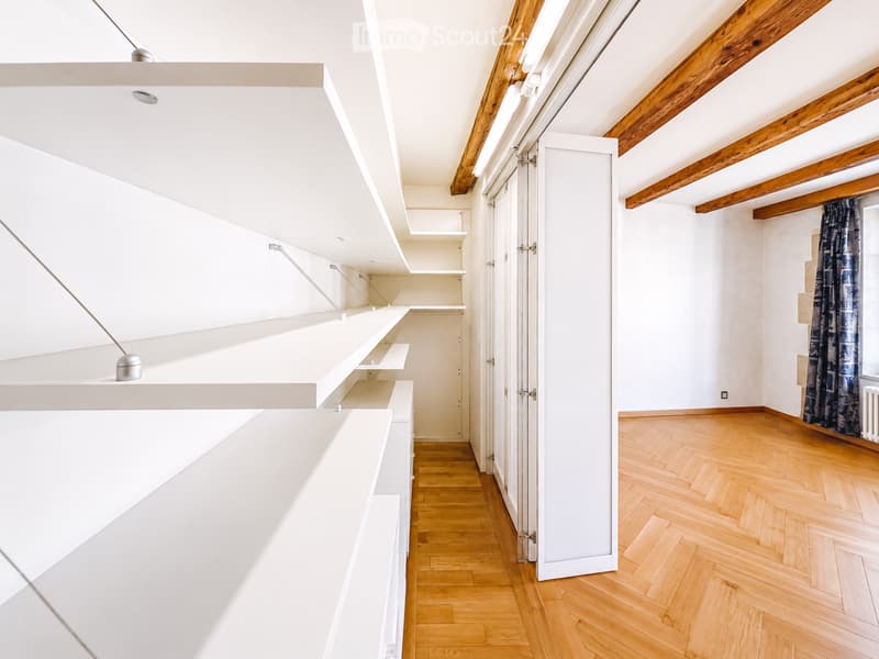 An unfurnished room with white built-in shelving units, wooden herringbone flooring, and exposed wooden beams on the ceiling. The room features a large window with a dark curtain and a radiator below it.
