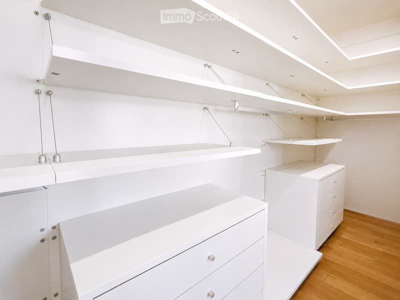 A modern, minimalist walk-in closet with white built-in shelving units, drawers, and a polished wooden floor.