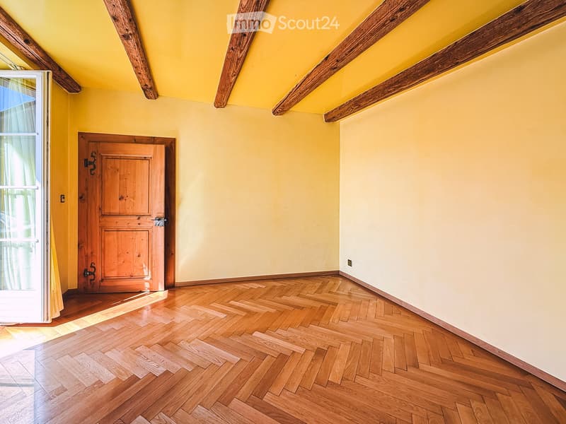 Empty room with wooden herringbone flooring, yellow walls, exposed wooden beams on the ceiling, and a wooden door.
