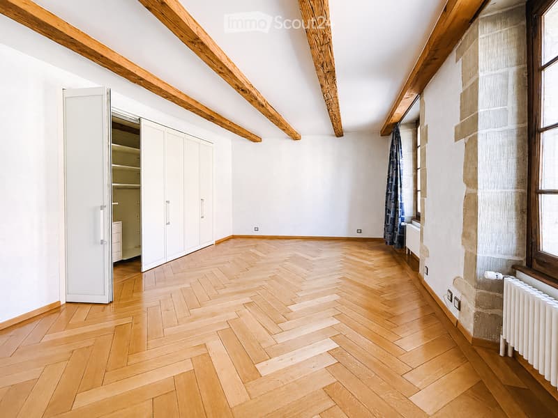 Empty room with wooden herringbone flooring, white walls, exposed wooden beams, and a large window with a curtain. A built-in closet with white doors is visible on the left side.