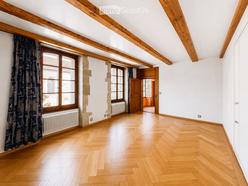 Empty room with wooden herringbone flooring, exposed wooden beams on the ceiling, large windows with wooden frames, and radiators beneath the windows.