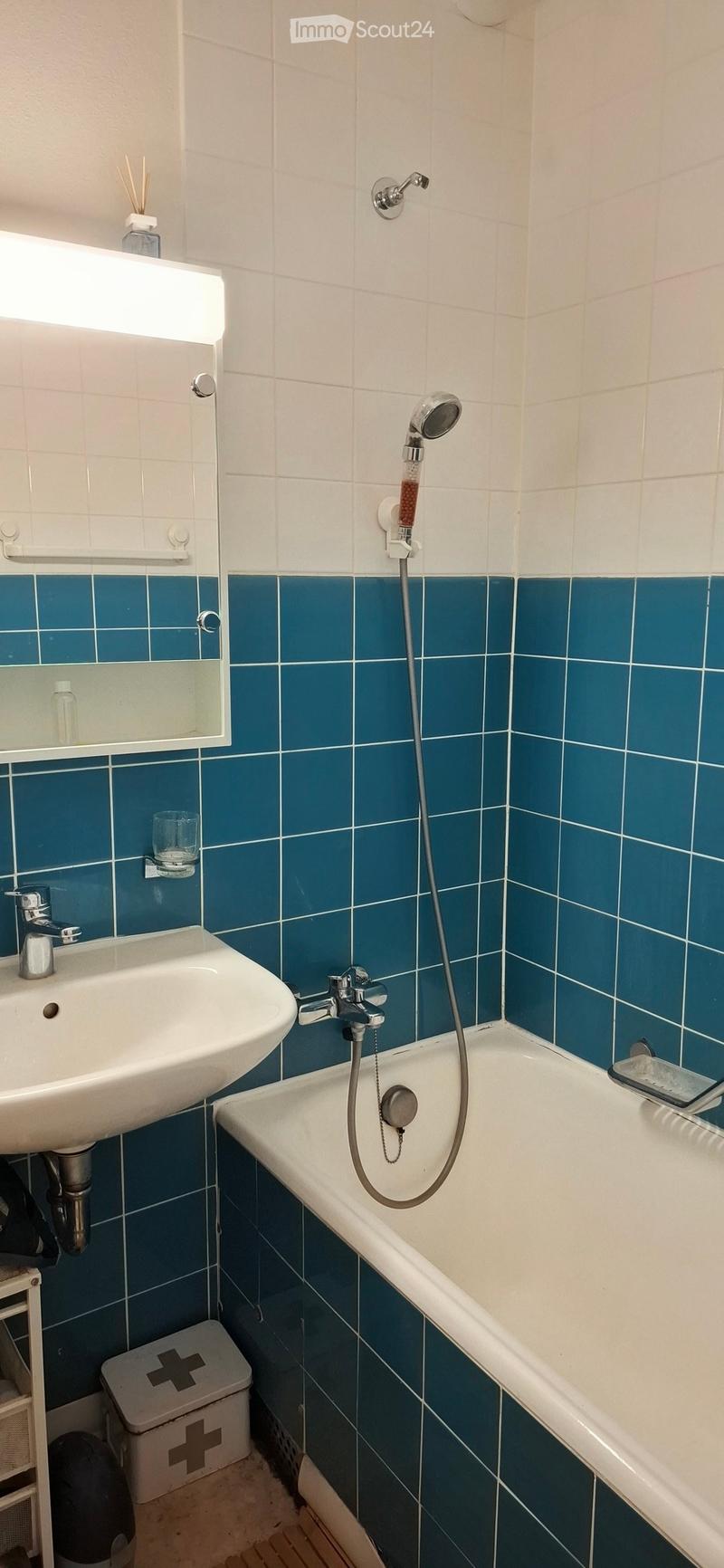 Bathroom with white tiles, blue lower tiles, a sink, bathtub, showerhead, mirror, and a small stool with a first aid kit.