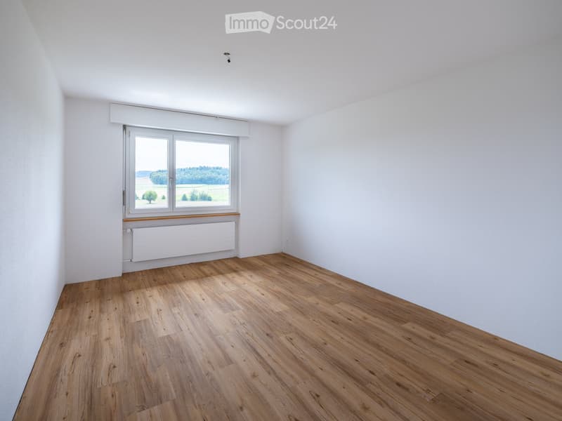 Empty room with wooden flooring, white walls, and a large window offering a view of a rural landscape.