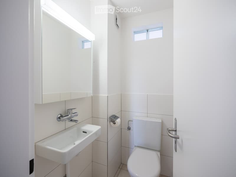 Modern, compact bathroom with white tiles, a single sink, a toilet, and a mirror above the sink. The room features a small window and a door with a silver handle.