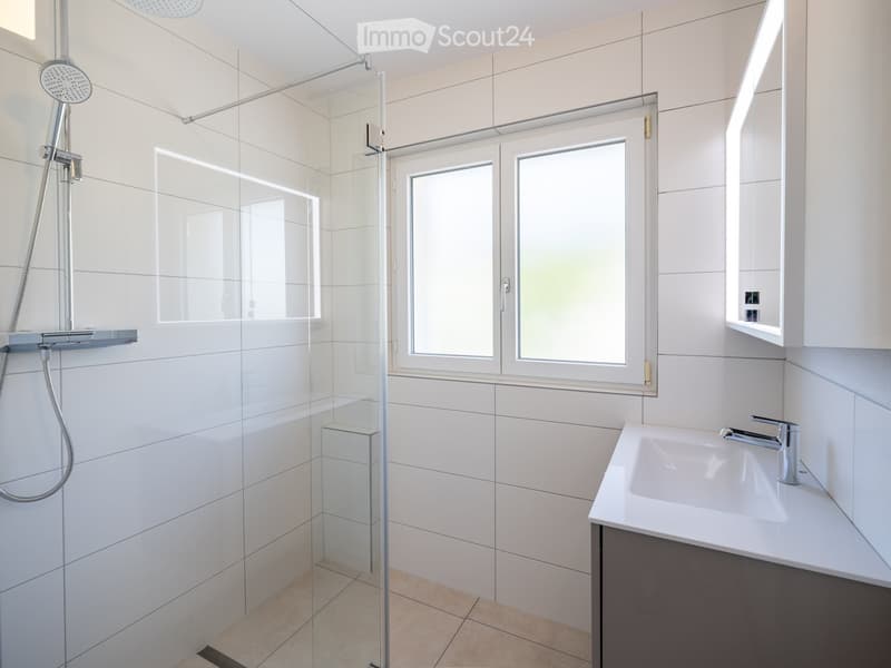 Modern bathroom with white tiles, glass-enclosed shower, and a white sink with a chrome faucet.