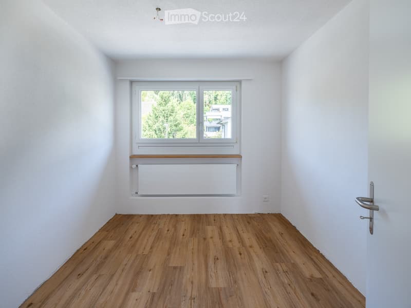 Empty room with wooden flooring, white walls, a window with a radiator below, and a door handle visible on the right side.
