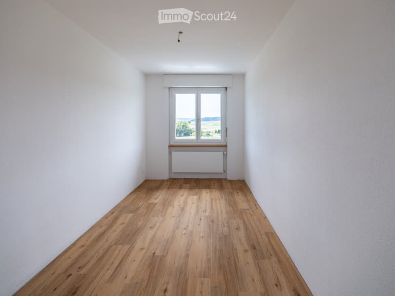 Empty room with wooden flooring, white walls, and a large window with a view of the landscape.